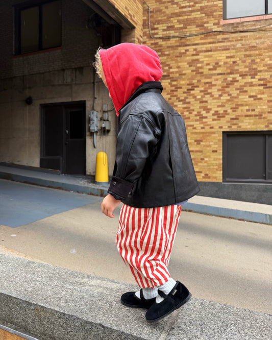Toddler wearing a red hoodie, black leather jacket, and red and white striped pants walking on a street.