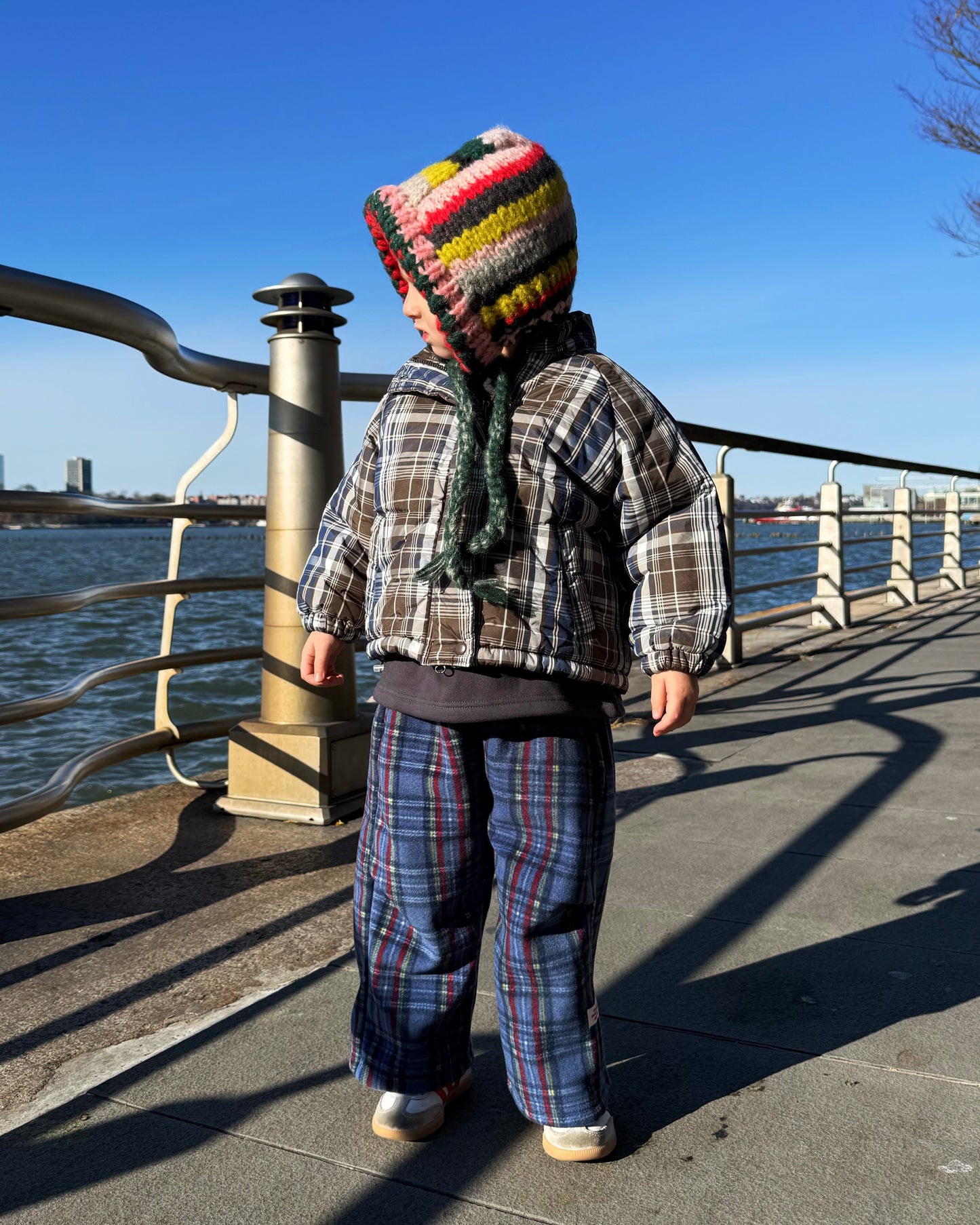 Child wearing a plaid puffer jacket and colorful knit hat standing by a waterfront with a clear blue sky.