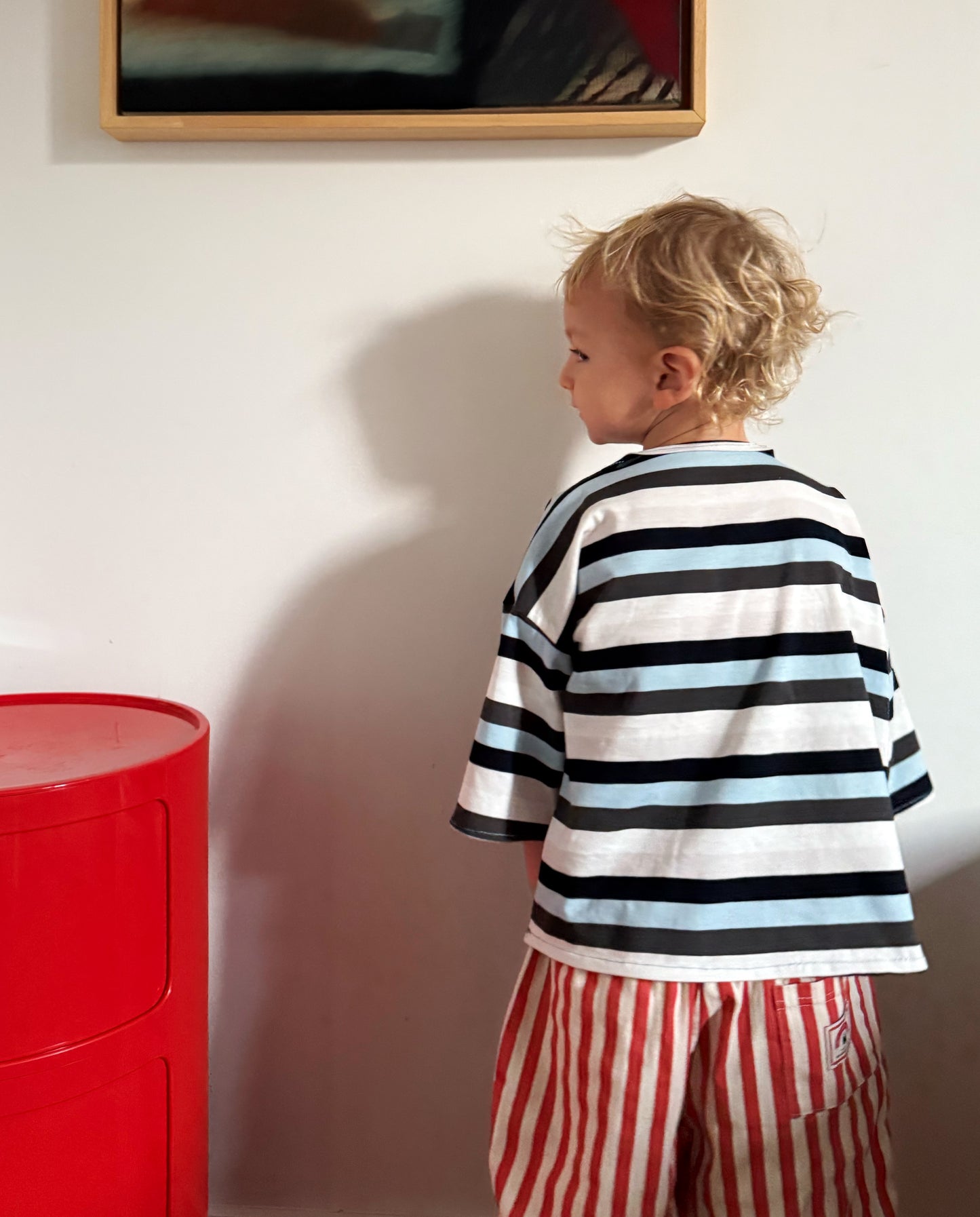 Child wearing a custom striped shirt and red pants standing against a white wall indoors. 