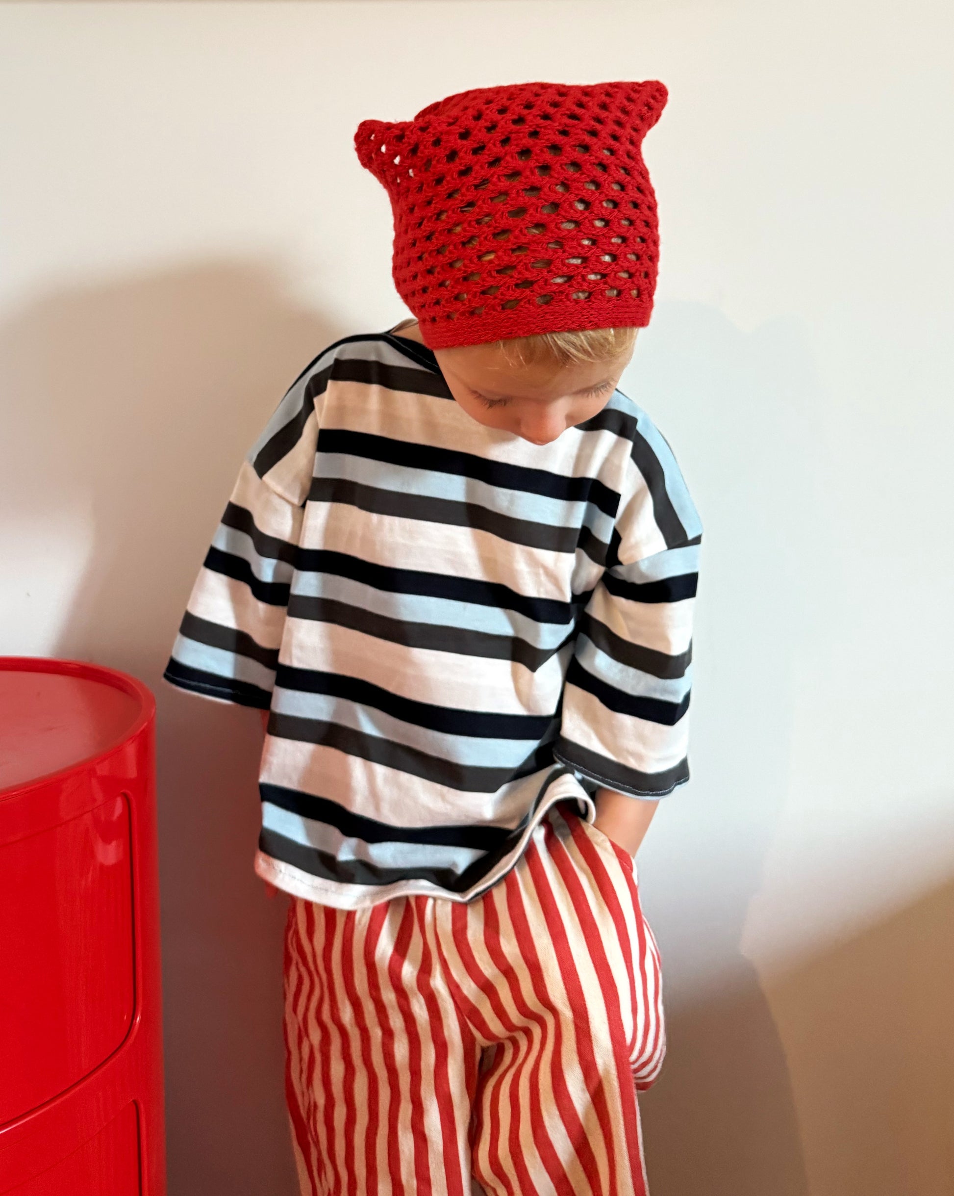 Child wearing a red knit hat, striped shirt, and red and white striped pants against a plain background.