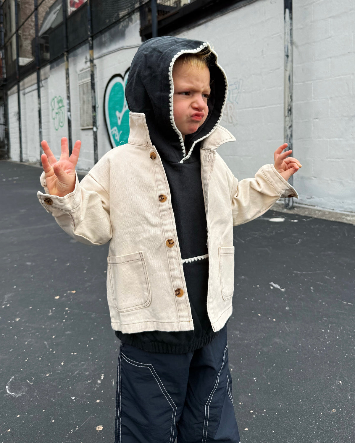 Child wearing a cream canvas jacket with brown buttons and a black hoodie outdoors.
