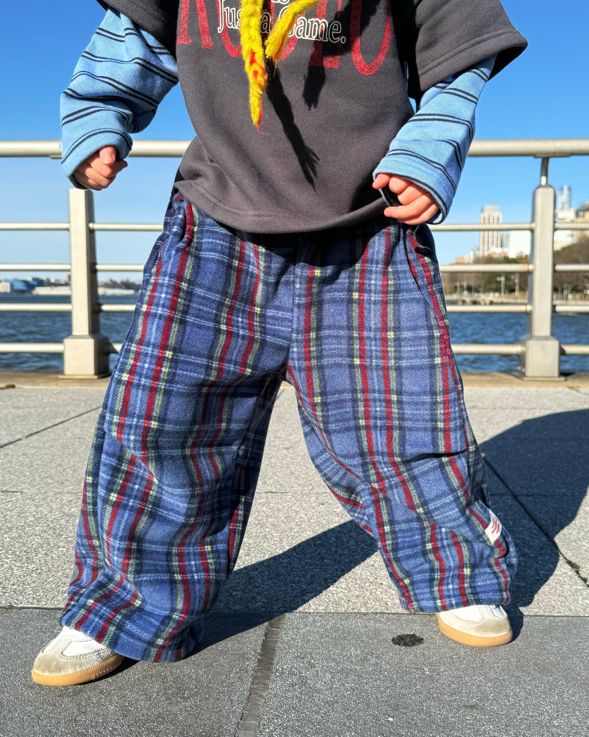 Kid wearing a gray and blue layered shirt with colorful text, cozy blue plaid pants, and white shoes by a waterfront.