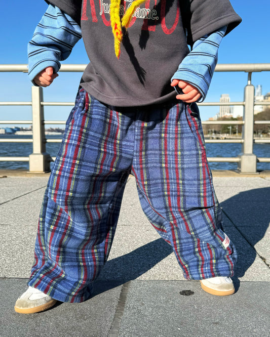 Kid wearing a gray and blue layered shirt with colorful text, cozy blue plaid pants, and white shoes by a waterfront.