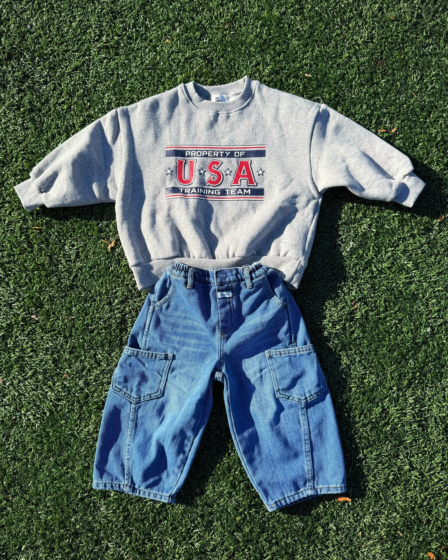 Gray toddler sweatshirt with 'Property of USA Training Team' text and blue jeans on grass.