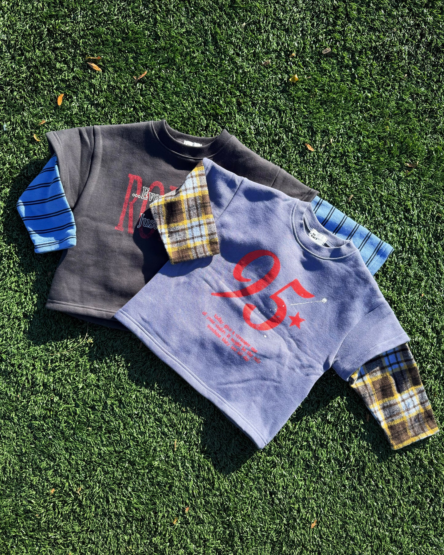 Two children's sweatshirts with plaid sleeves on a grassy surface
