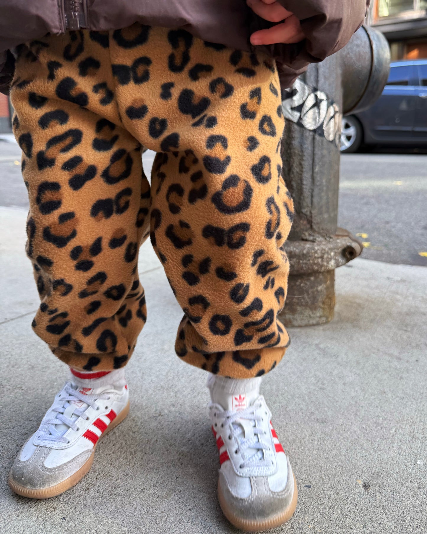 Toddler wearing leopard print fleece pants and white sneakers with red accents on a sidewalk.