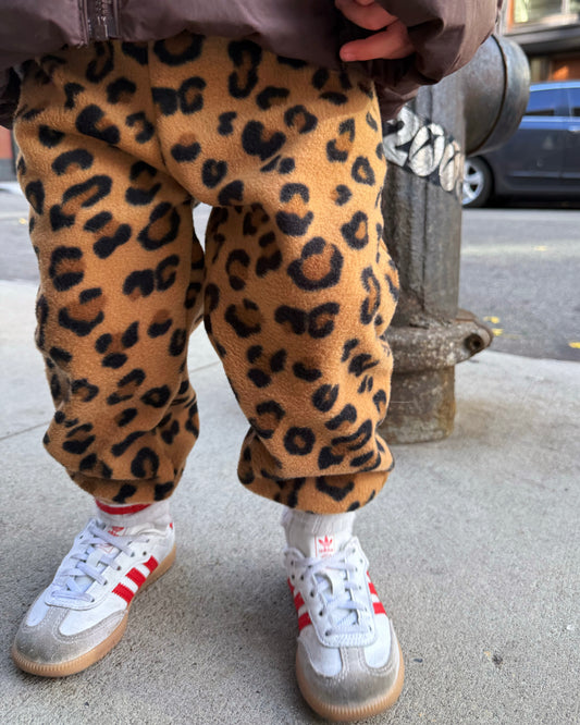 Toddler wearing leopard print fleece pants and white sneakers with red accents on a sidewalk.