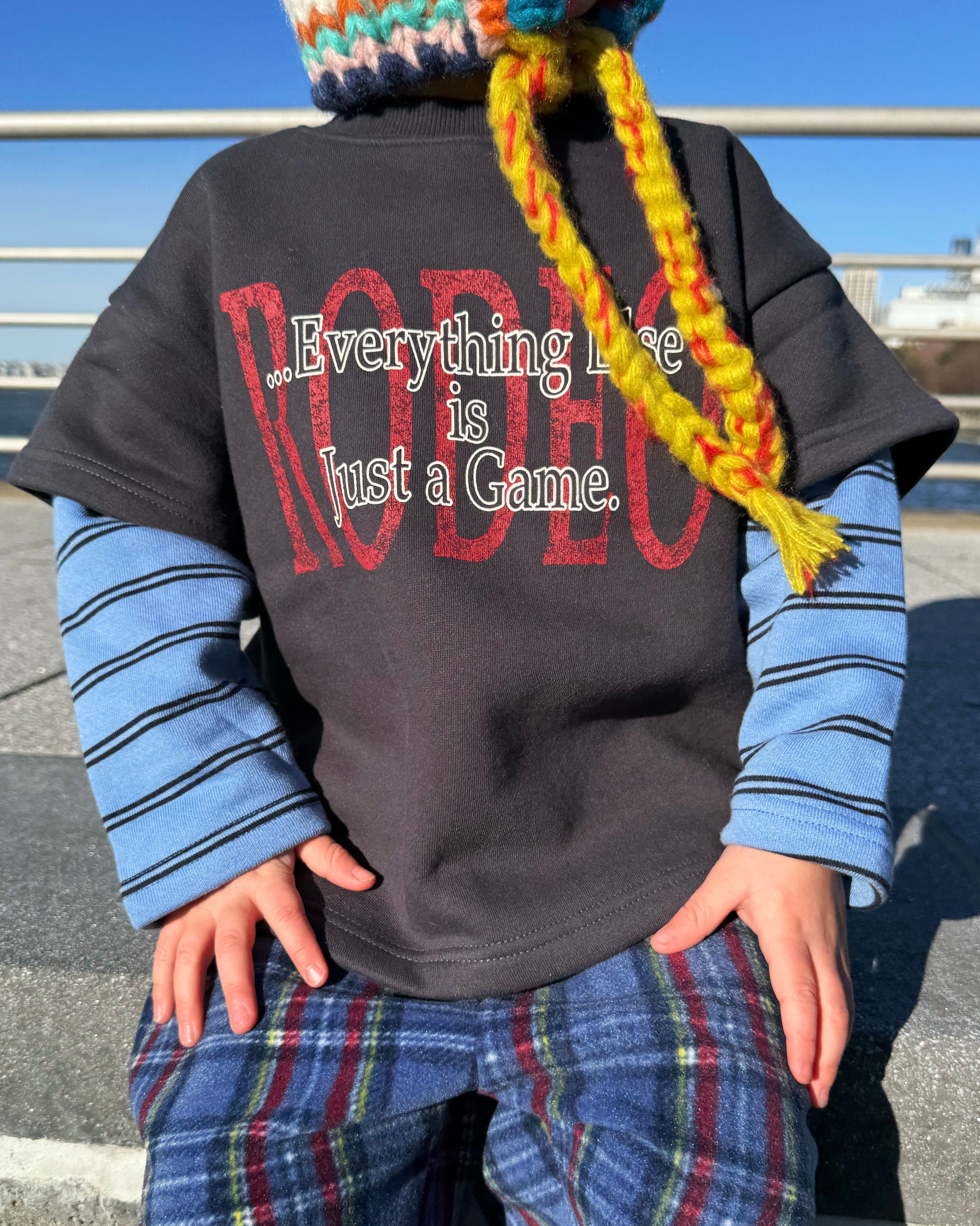 Kid wearing a black t-shirt with text, blue striped sleeves, and plaid pants outdoors.