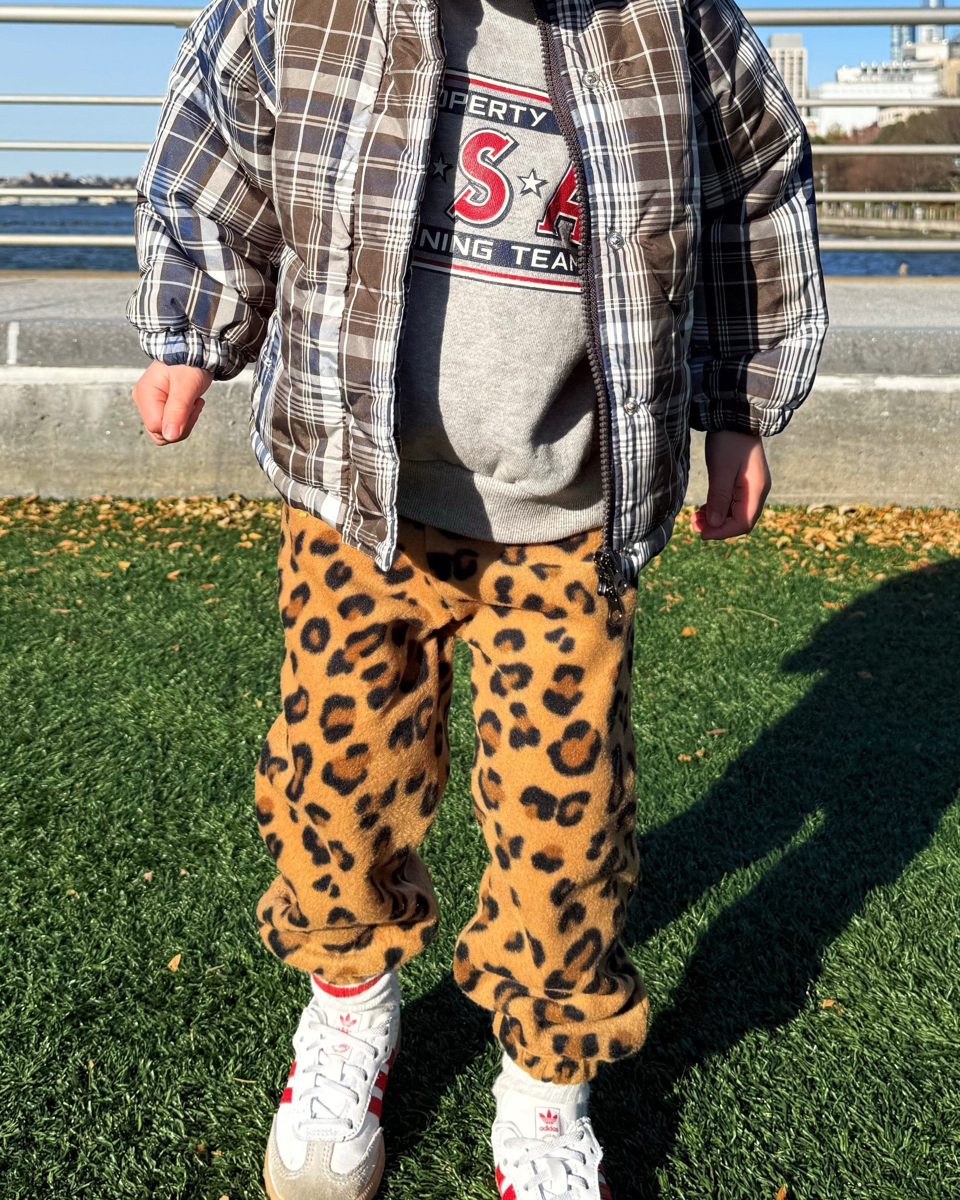 Toddler wearing a plaid padded jacket, gray sweatshirt with red text, and cozy leopard print fleece pants outdoors.