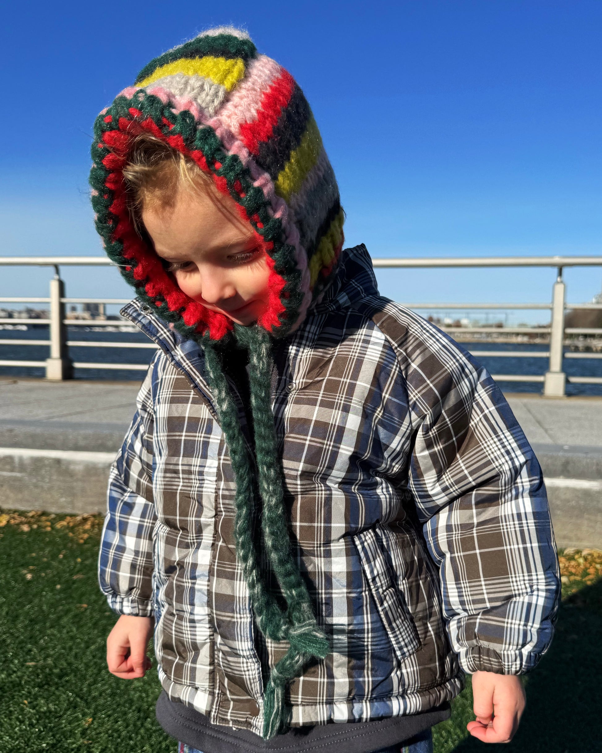 Child wearing a colorful knitted hat and plaid jacket outdoors on a clear day.