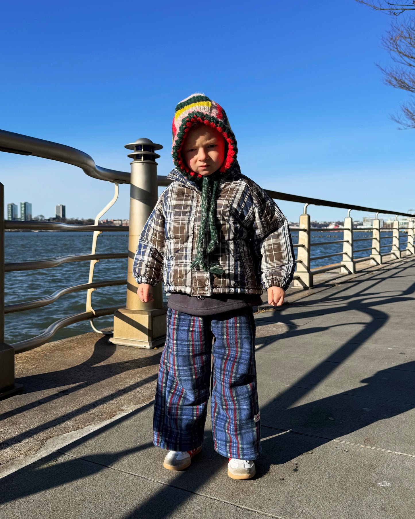 Child in plaid outfit and colorful knit hat standing by a waterfront on a clear day
