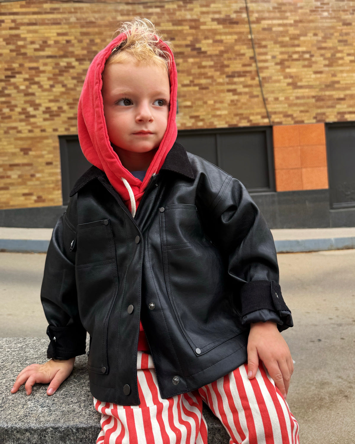 Child wearing a black leather jacket over a red hoodie with striped pants against a brick wall.